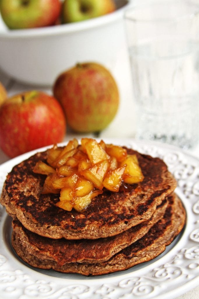 Pilha com 3 panquecas de maçã com cobertura de maçã caramelizada sobre prato branco em uma mesa branca