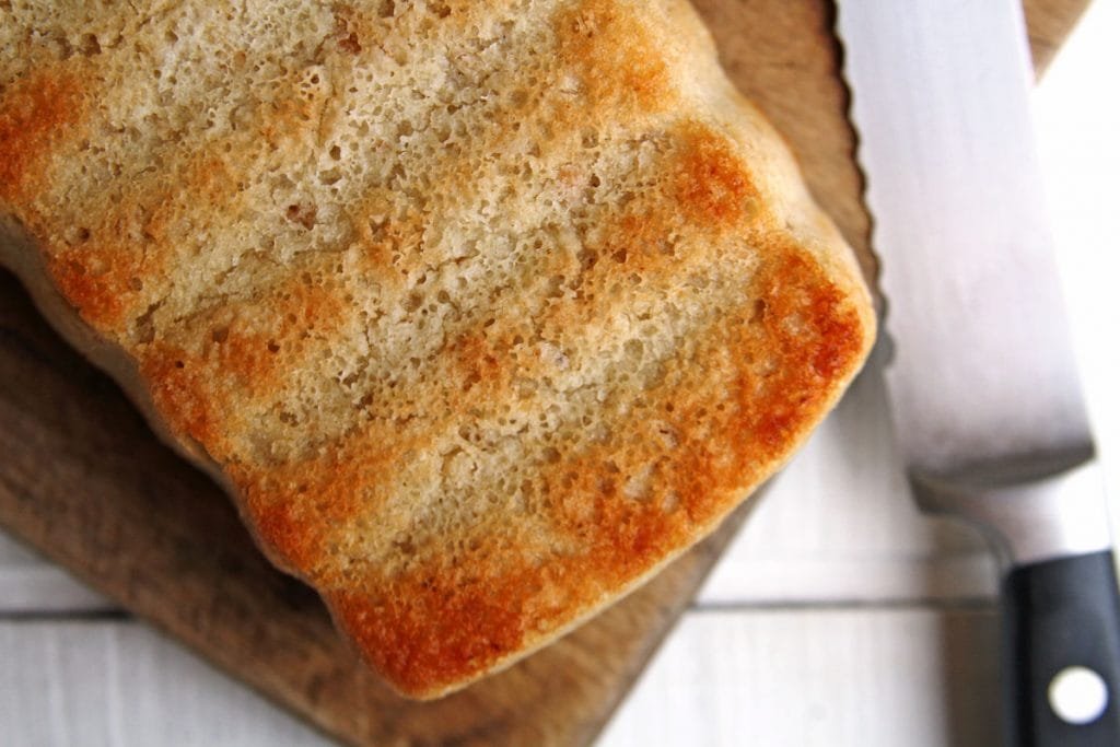 Pão sobre tábua de madeira com uma faca ao lado em uma mesa branca