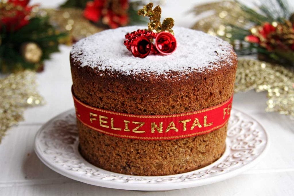 Panetone sobre prato branco, decorado com fita vermelha, açúcar confeiteiro e enfeites de natal