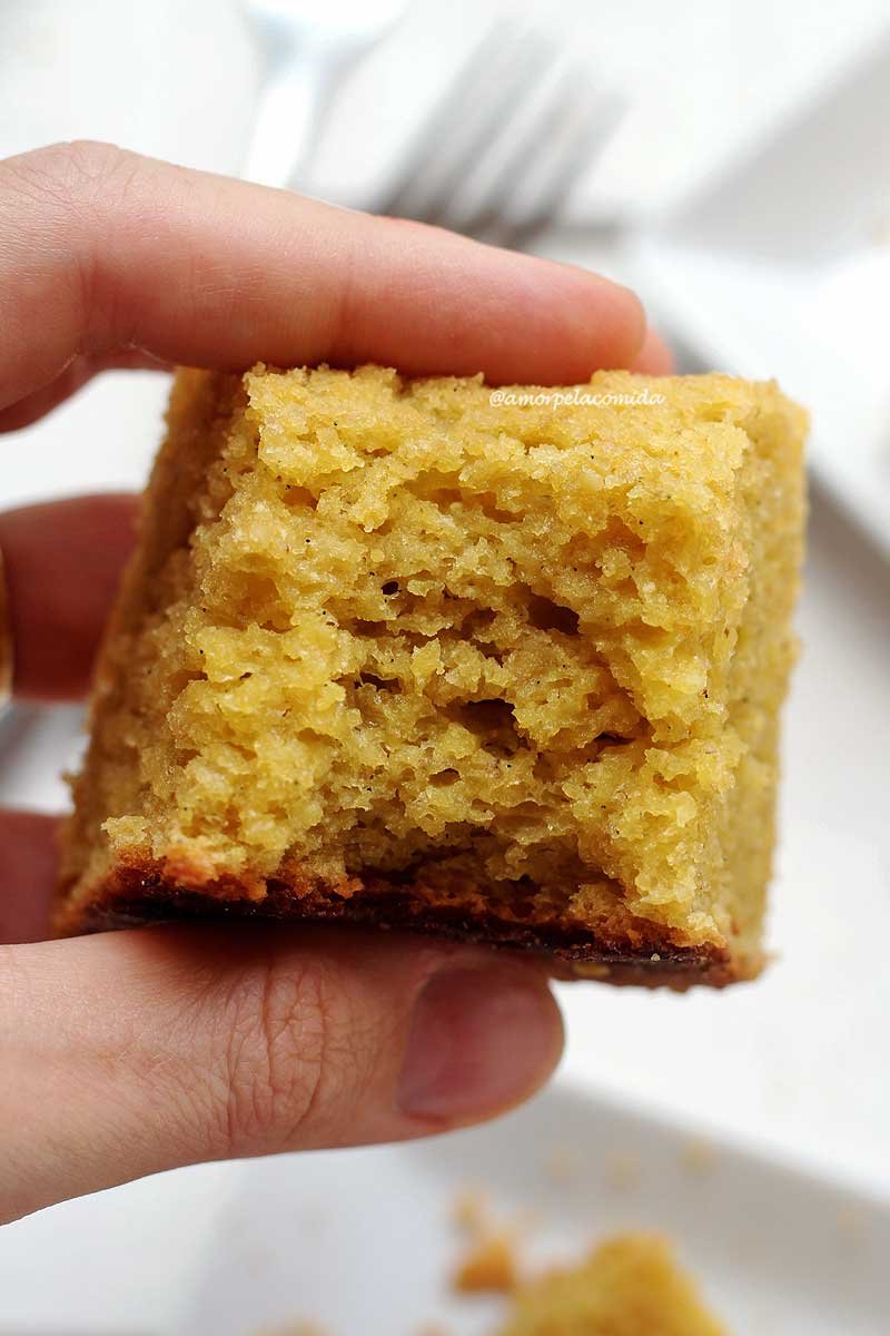 Pedaço de bolo de coco mordido sendo segurado por uma mão sobre uma mesa branca