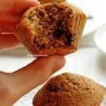 Mão segurando bolo de baunilha com gotas de chocolate pequeno mordido sobre mesa branca com outro bolinho pequeno sobre prato redondo branco