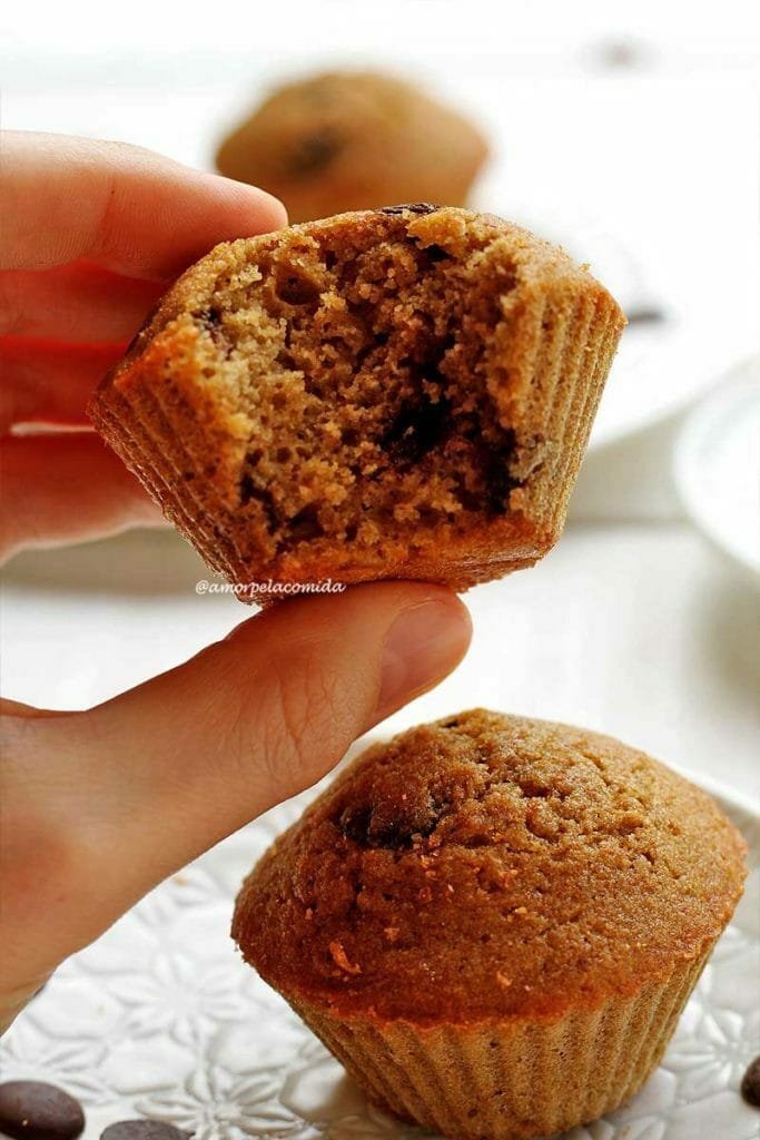 Mão segurando bolo de baunilha com gotas de chocolate pequeno mordido sobre mesa branca com outro bolinho pequeno sobre prato redondo branco