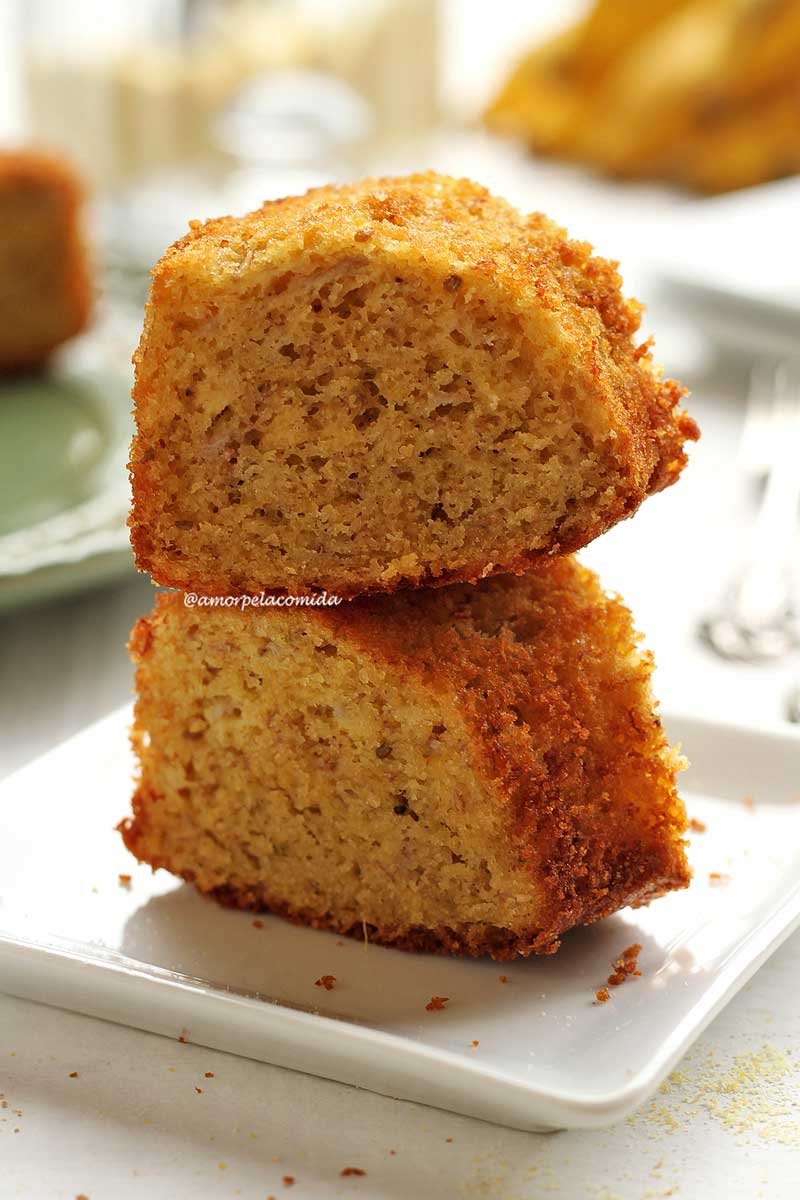 Duas fatias de bolo de fubá sobrepostas em um prato branco quadrado pequeno em mesa branca