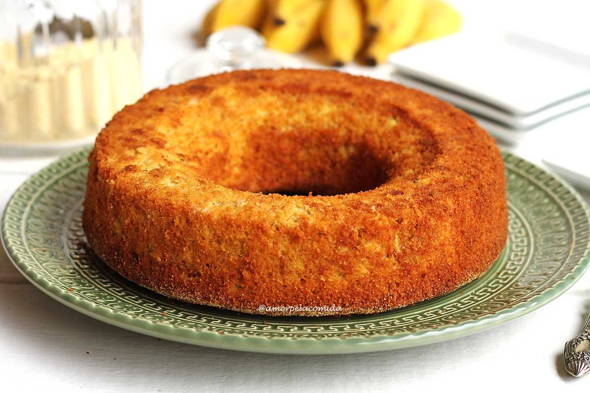 Bolo de fubá redondo com furo no meio sobre prato verde em uma mesa branca com bananas ao fundo