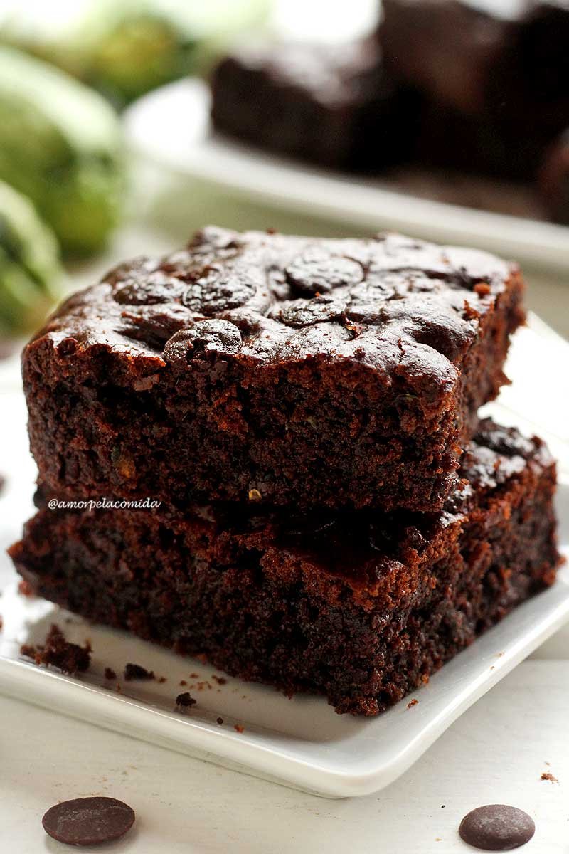 Dois pedaços de brownie sobrepostos em um prato quadrado branco em mesa branca ao fundo duas abobrinhas e mais pedaços de brownie em outro prato