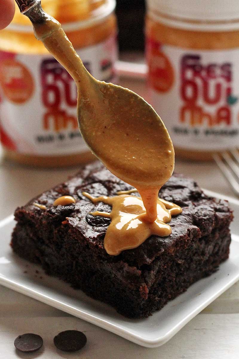 Pedaço de brownie sobre prato quadrado pequeno branco com manteiga de amendoim sendo derramada em cima com uma colher