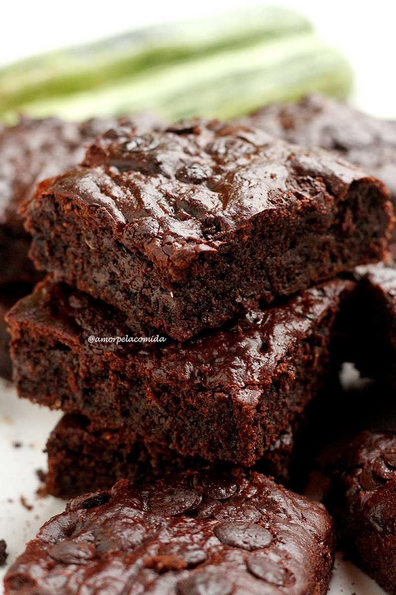 Vários pedaços de brownie sobrepostos em um prato branco, com abobrinhas bem ao fundo desfocadas