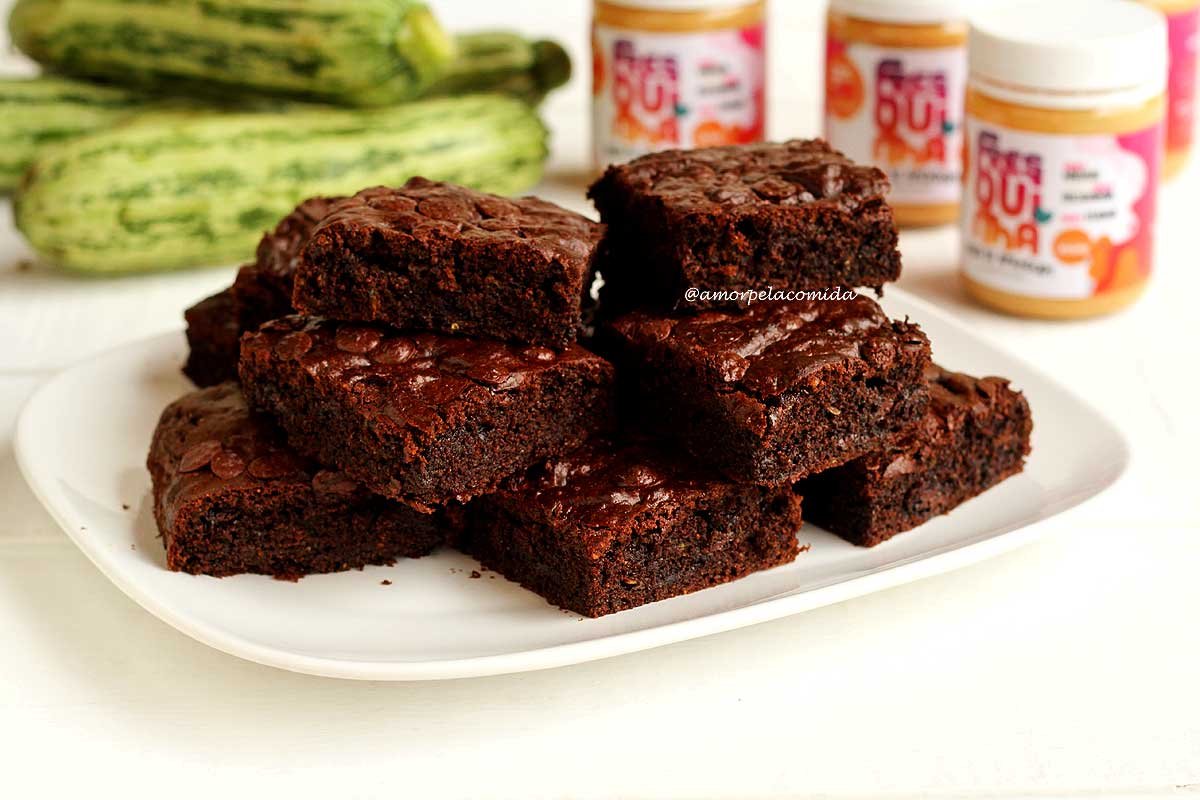 Prato branco quadrado com vários pedaços de brownie sobrepostos, ao fundo abobrinhas e potes de manteiga de amendoim