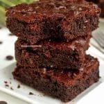 Três pedaços de brownie sobrepostos em um prato quadrado branco pequeno, o pedaço de cima está mordido e tem abobrinha ao fundo desfocadas