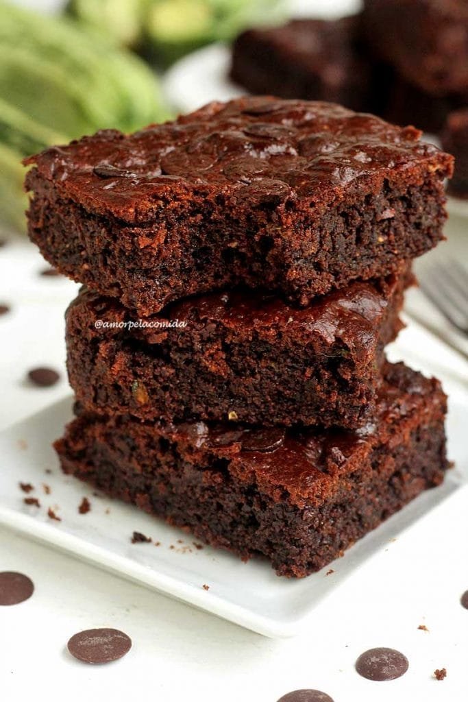 Três pedaços de brownie sobrepostos em um prato quadrado branco pequeno, o pedaço de cima está mordido e tem abobrinha ao fundo desfocadas