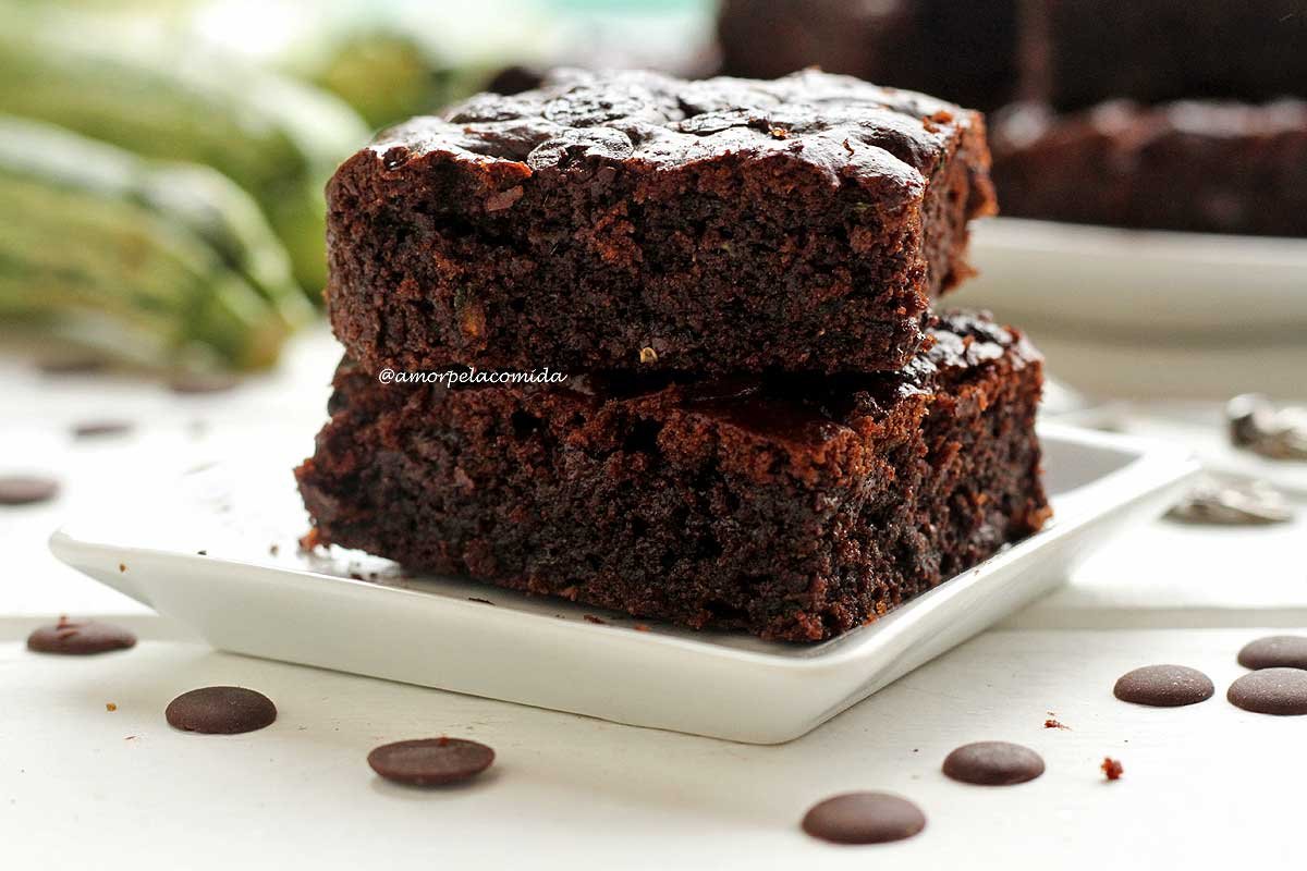 Duas fatias de brownie sobrepostas em um prato quadrado branco em uma mesa branca co abobrinhas ao fundo e mais pedaços de brownie desfocados