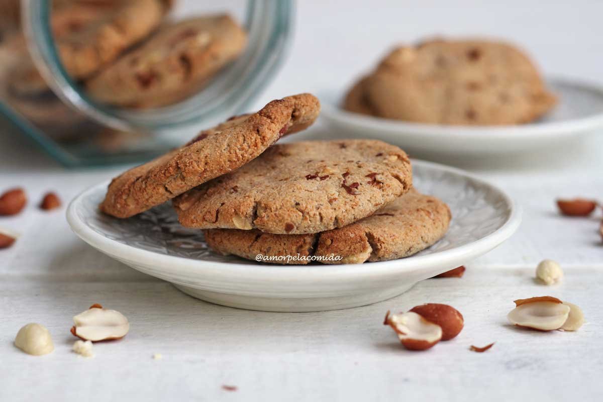 Três biscoitos de amendoim amontoados sobre prato branco redondo pequeno em mesa branca, ao fundo vidro com biscoitos deitado e mais um prato redondo com biscoitos