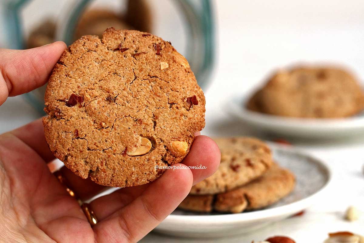 Mão segurando biscoito de amendoim sobre mesa branca, ao fundo prato pequeno com biscoitos