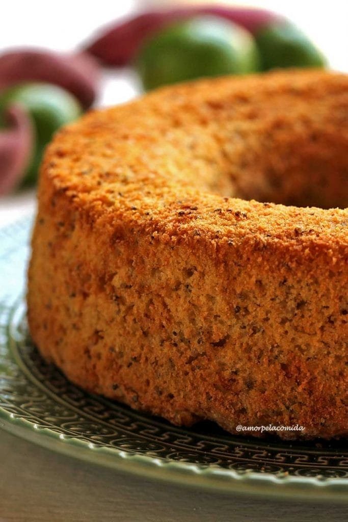 Bolo de limão redondo com furo no meio sobre prato verde escuro em uma mesa branca com batata-doce e limões ao fundo