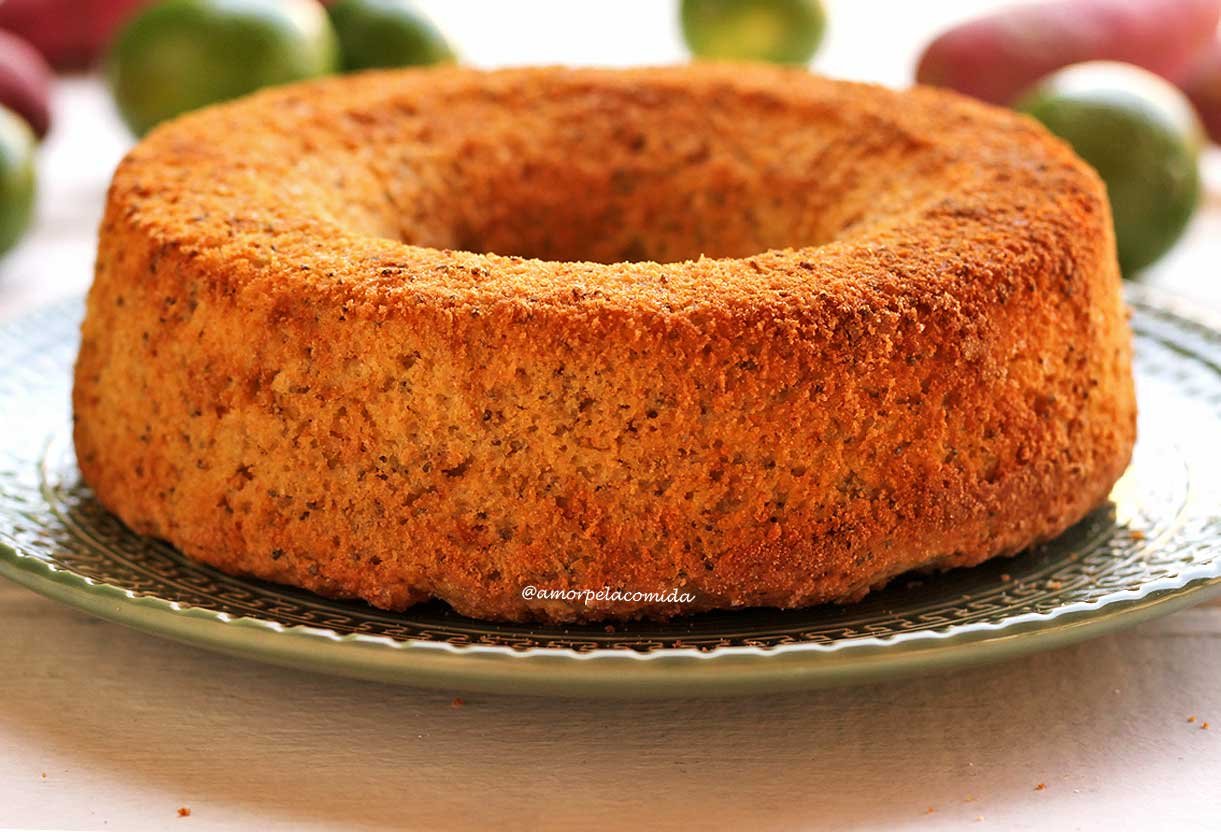 Bolo de limão redondo com furo no meio sobre um prato verde escuro em uma mesa branca com limões ao fundo