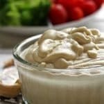 Pote de requeijão cremoso sobre um prato pequeno de prata com castanha de caju em volta e ao fundo vegetais em um prato branco