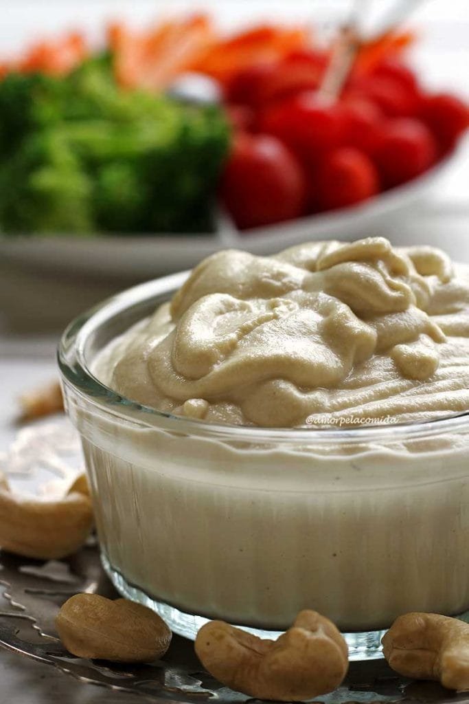 Pote de requeijão cremoso sobre um prato pequeno de prata com castanha de caju em volta e ao fundo vegetais em um prato branco