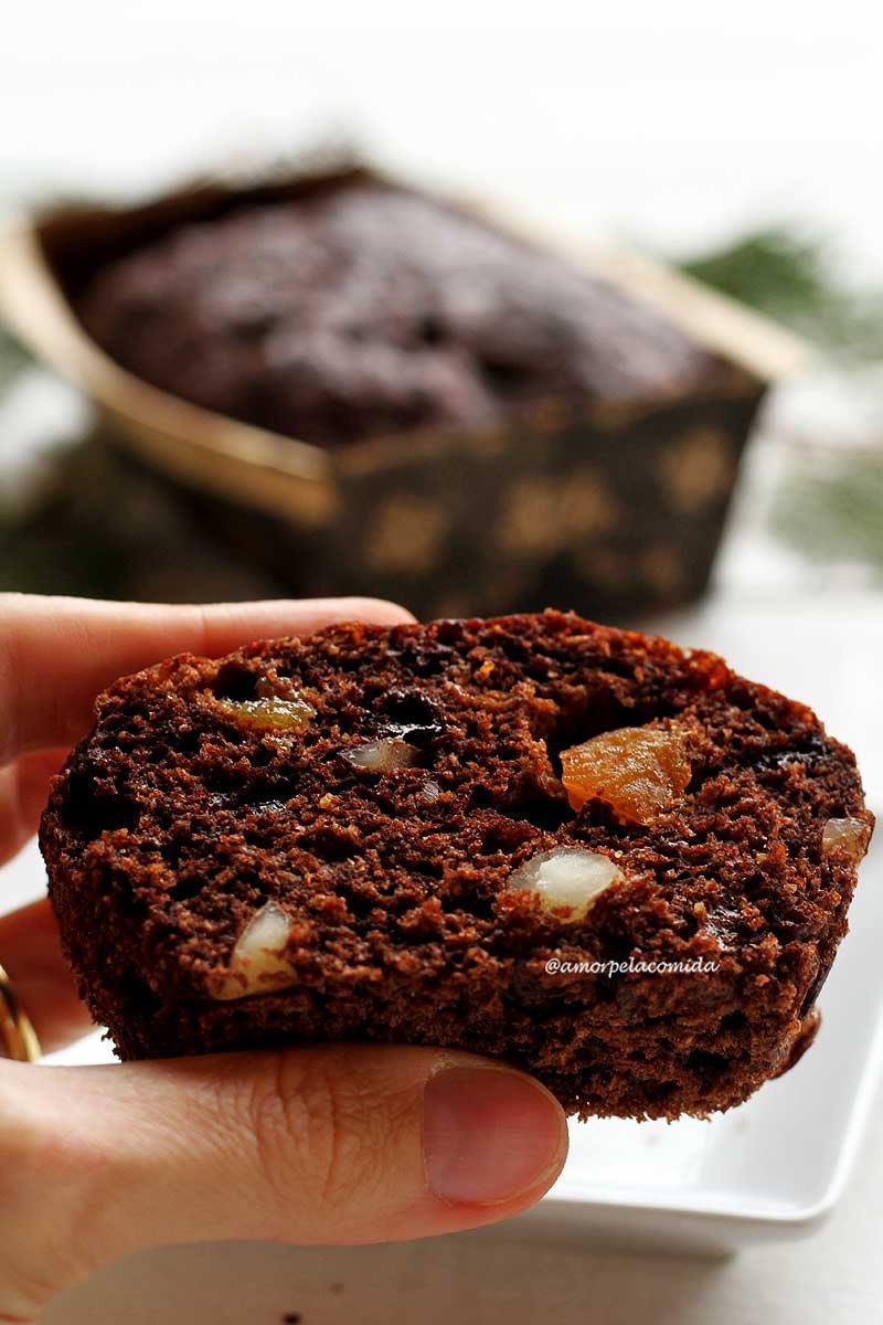 Mão segurando fatia de chocotone com castanhas e damasco no meio, ao fundo forma onde foi assado o chocotone