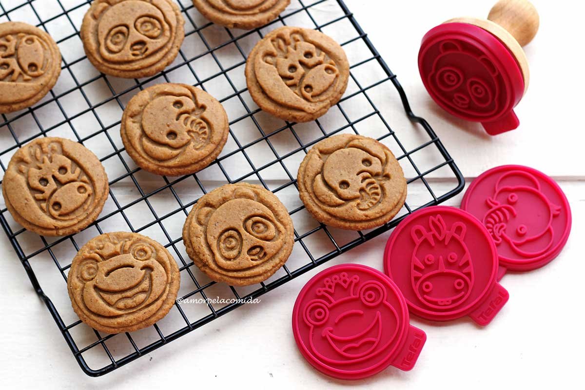 Black grid with cookies with faces cooling on a white table, next to the stamp that allows the cookies to be embossed