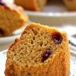 Uma fatia de bolo de fubá com pedaços de goiabada sobre prato quadrado branco, ao fundo mais uma fatia cortada e o bolo desfocado
