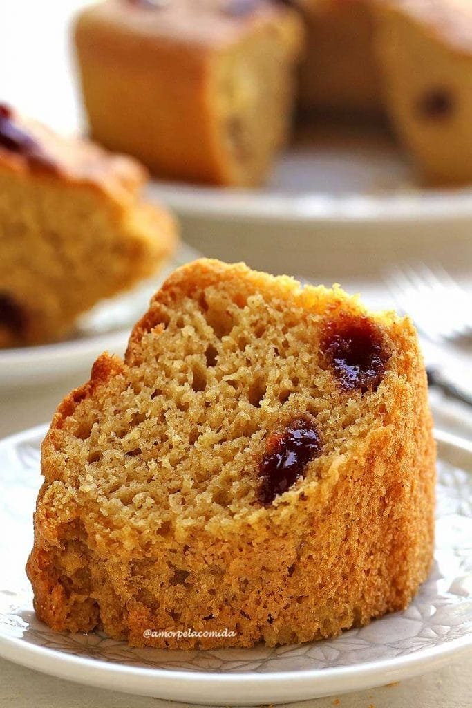 Uma fatia de bolo de fubá com pedaços de goiabada sobre prato quadrado branco, ao fundo mais uma fatia cortada e o bolo desfocado