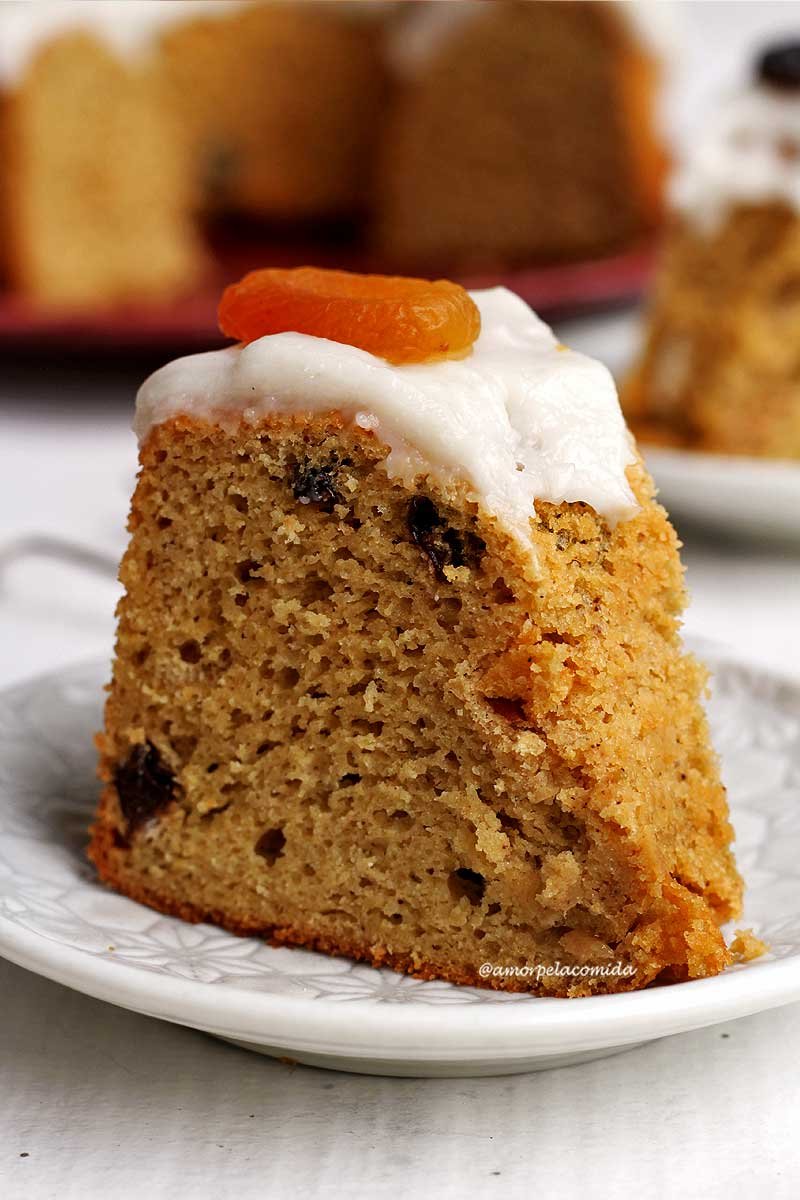 Pedaço de bolo de frutas com cobertura branca decorado com um damasco por cima sobre um prato redondo branco pequeno