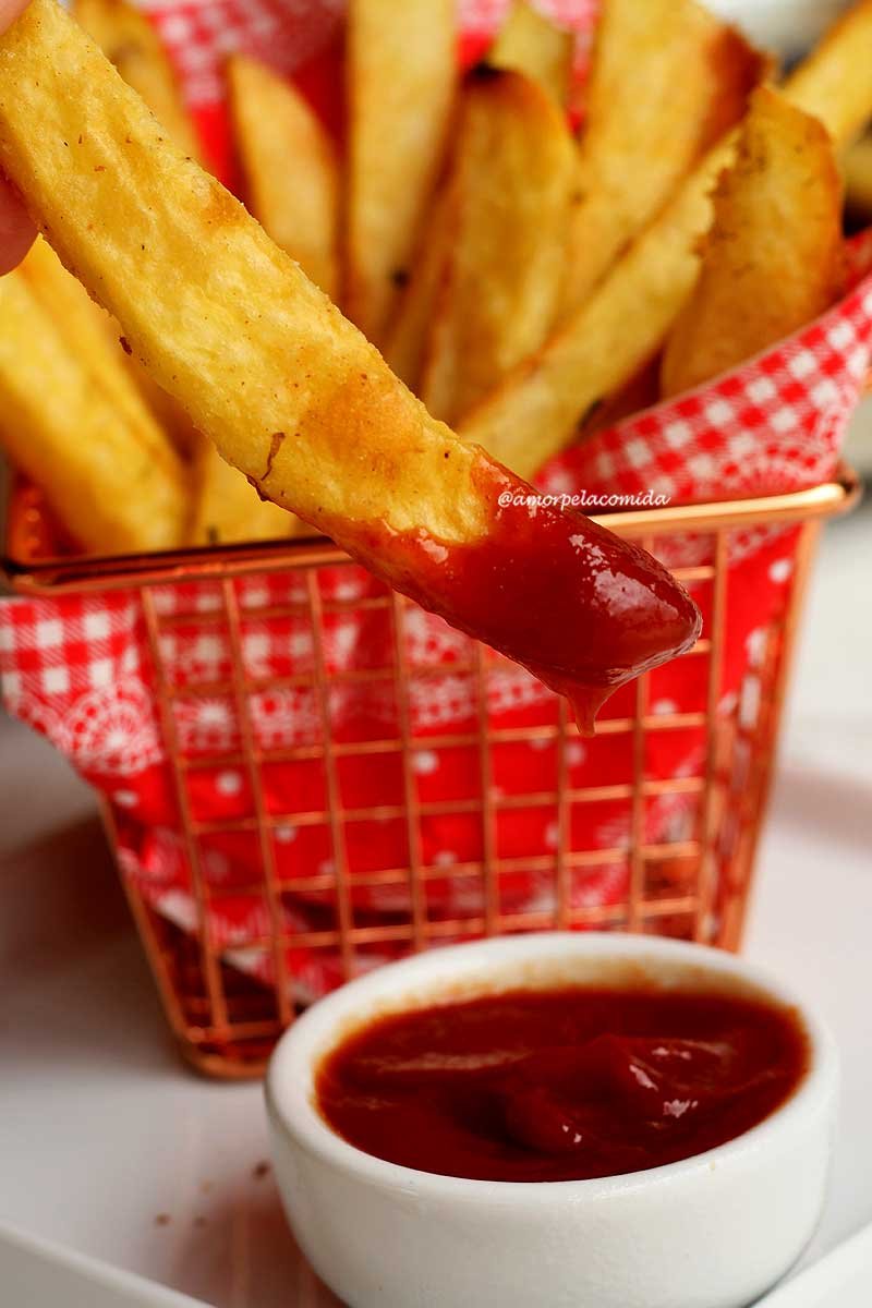 Mão segurando tolete de batata assada, a ponta do tolete foi mergulhada no ketchup, o pote de ketchup está logo abaixo do tolete de batata, atrás cestinha de metal com várias batatinhas 