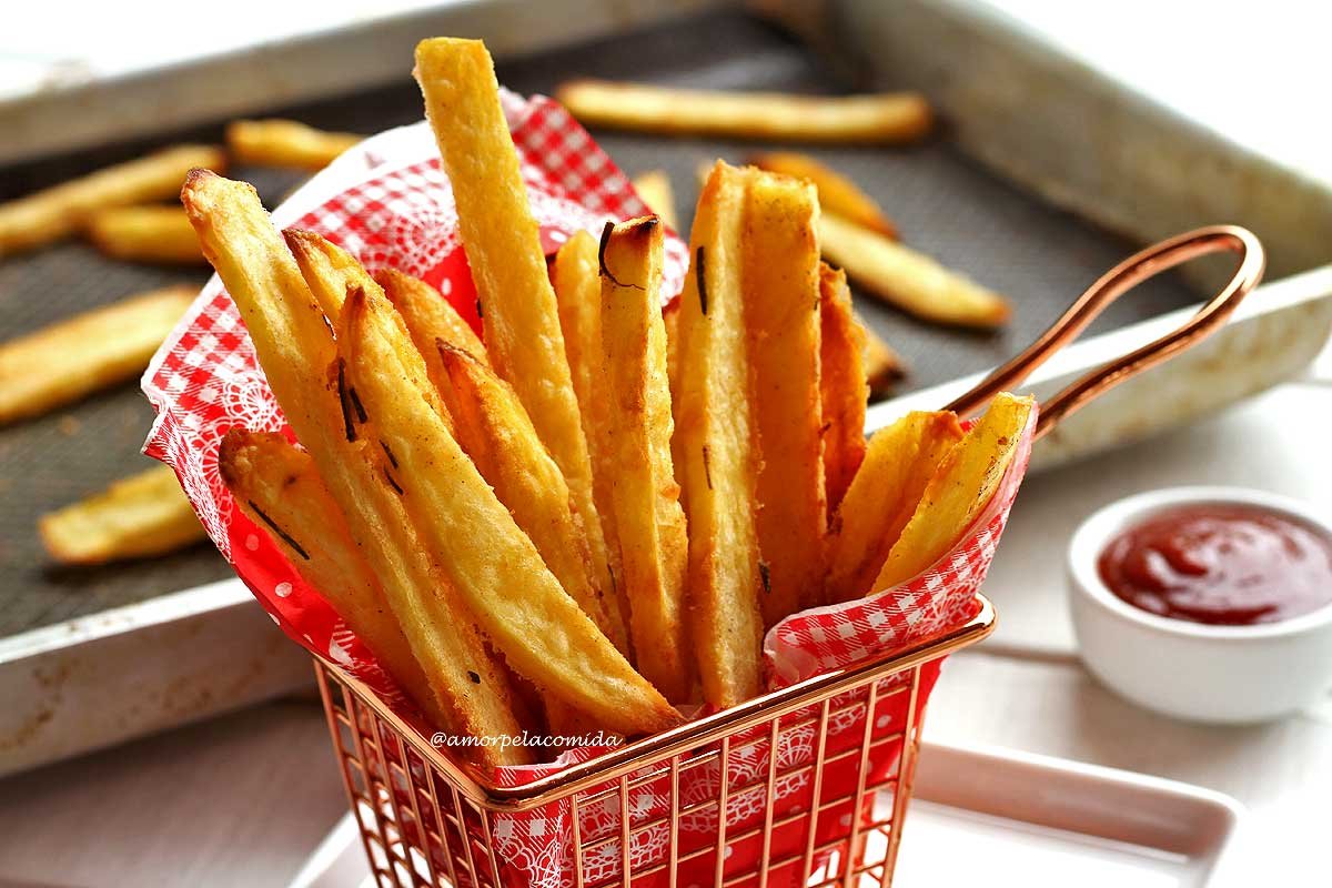 Small metal basket with a red napkin with white checkerboard, inside several pieces of oven-baked French fries, next to the basket a small jar of ketchup, in the background the pan where the potatoes were baked