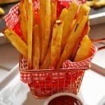 Cestinha de metal pequena com guardanapo vermelho com xadrez branco, dentro vários toletes de batata frita assada no forno, ao lado da cestinha um pote pequeno com ketchup, ao fundo a forma onde foram assadas as batatas