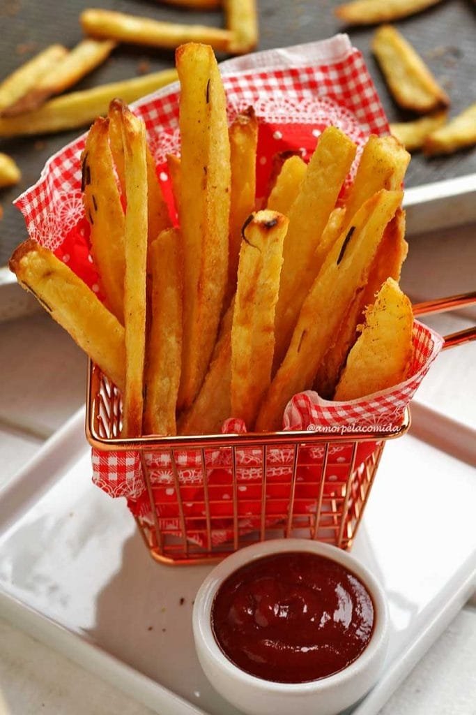 Small metal basket with a red napkin with white checkerboard, inside several pieces of oven-baked French fries, next to the basket a small jar of ketchup, in the background the pan where the potatoes were baked