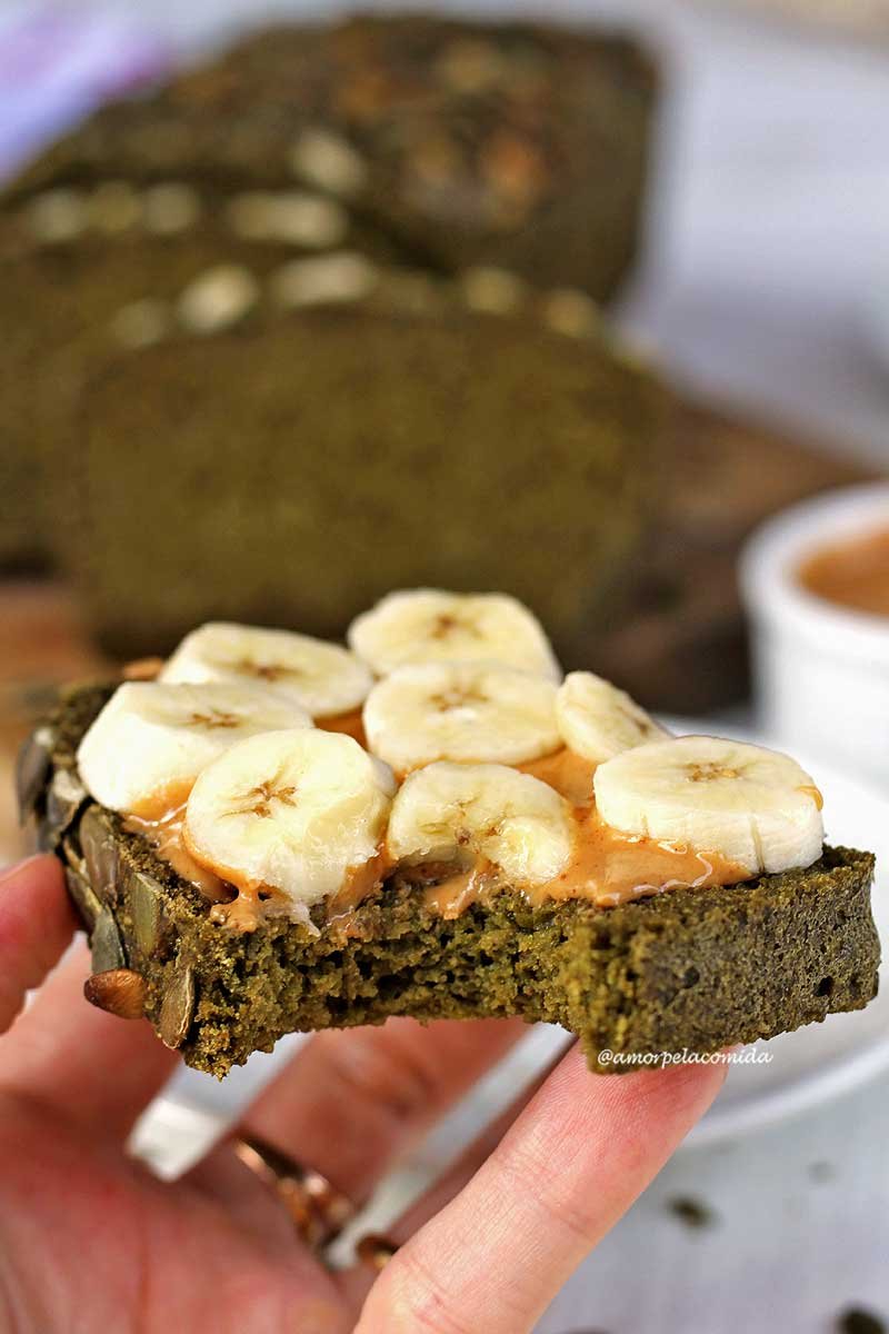 Mão segurando fatia de pão mordido feito a partir da receita com ora-pro-nóbis. A fatia está coberta com uma camada de pasta de amendoim e fatias de banana, ao fundo o pão de forma desfocado sobre a tábua de madeira