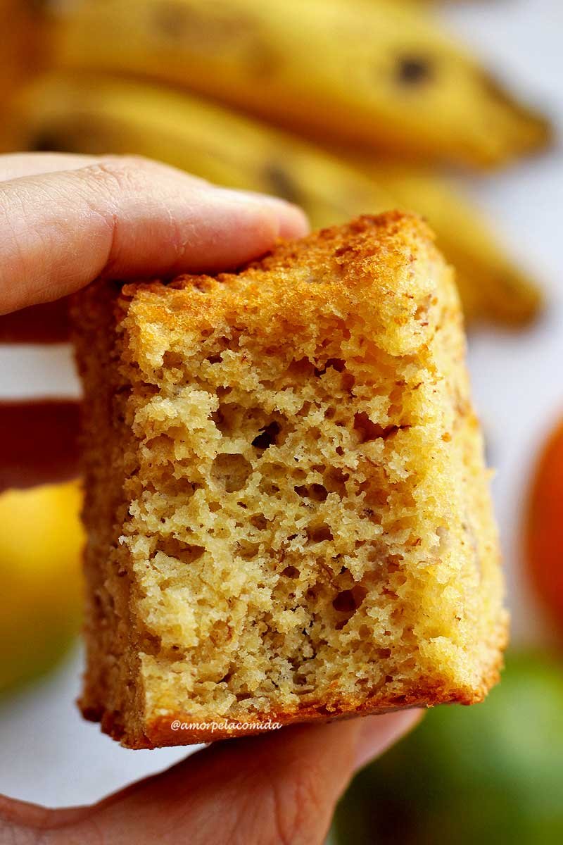 Pedaço de bolo de fubá com laranja e banana mordido sendo segurado por uma mão, ao fundo bananas e laranjas desfocadas em uma mesa branca