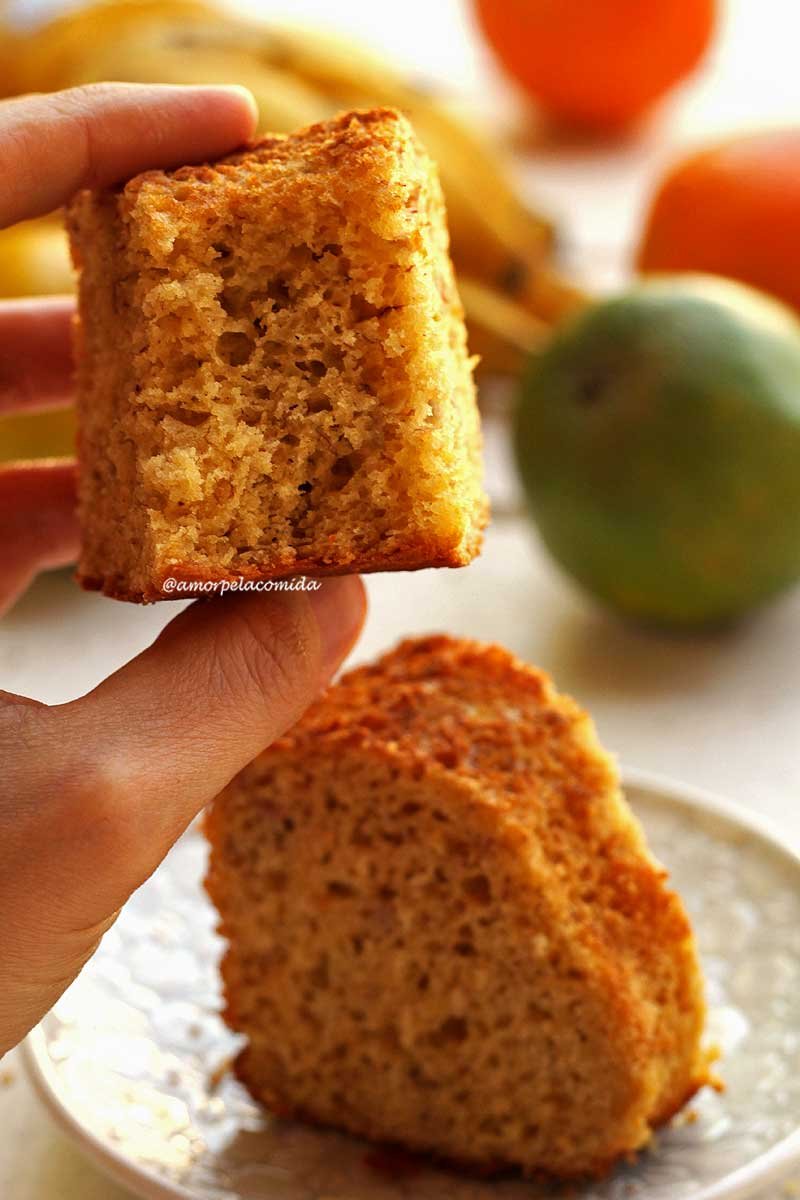 Mão segurando pedaço mordido de bolo de fubá com laranja e banana, abaixo uma fatia de bolo sobre prato redondo pequeno em uma mesa branca, ao fundo laranjas e bananas desfocadas