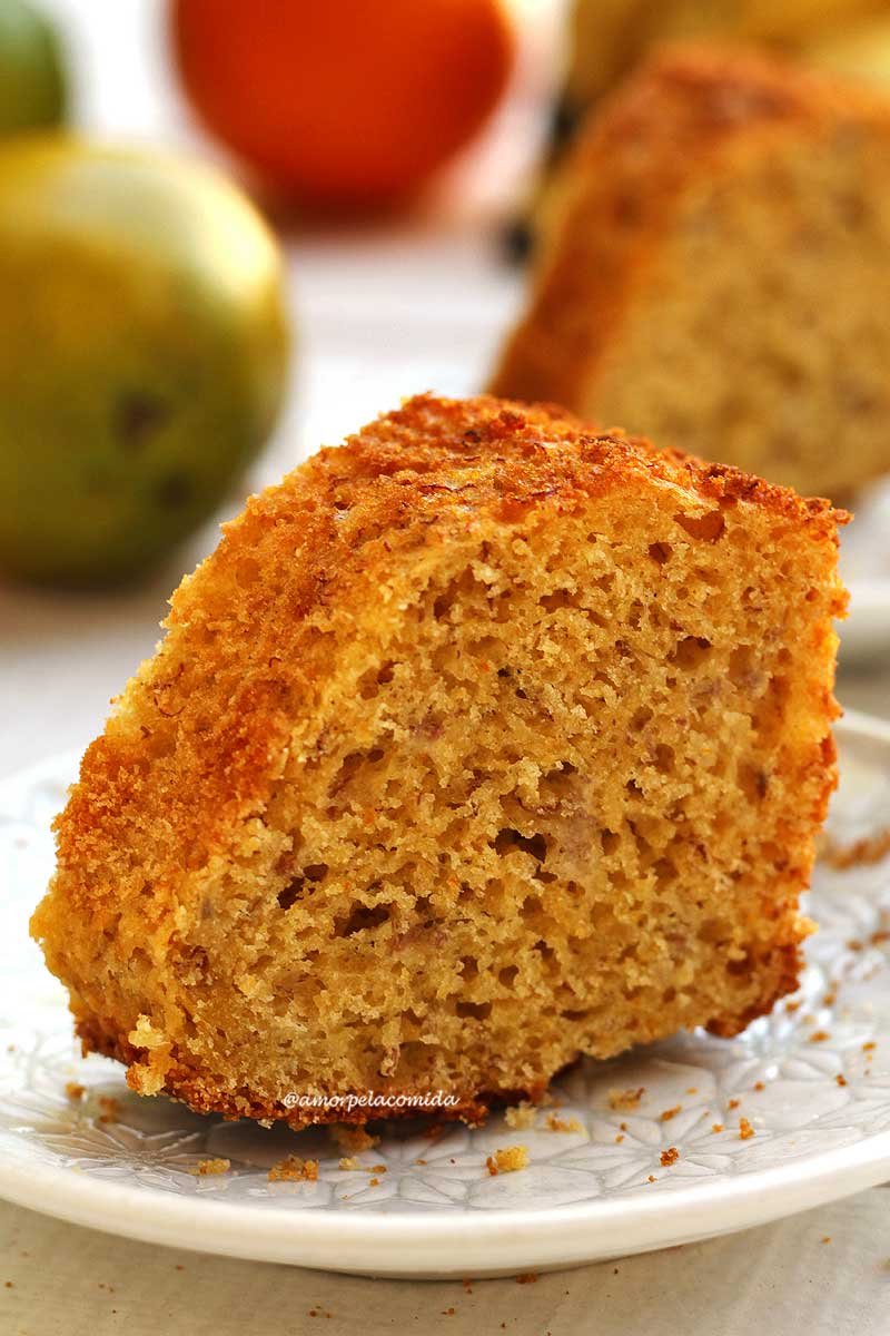 Fatia de bolo de fubá com laranja e banana sobre prato branco pequeno em uma mesa branca, ao fundo laranjas e outra fatia de bolo desfocada