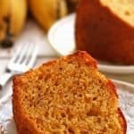 Fatia de bolo de fubá com laranja e banana deitado dobre prato redondo branco pequeno, ao fundo mais uma fatia de bolo e bananas desfocadas