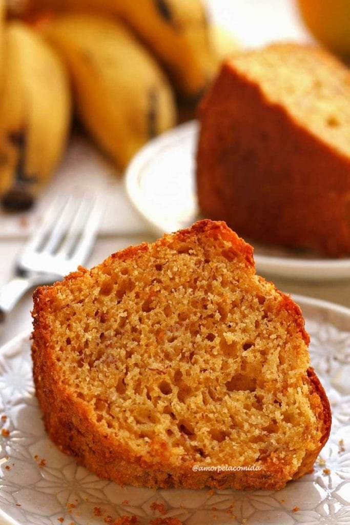 Fatia de bolo de fubá com laranja e banana deitado dobre prato redondo branco pequeno, ao fundo mais uma fatia de bolo e bananas desfocadas