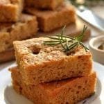 Dois pedaços quadrados de focaccia sobre prato branco, ao fundo um pote de azeite pequeno e vários pedaços cortados de focaccia sobre uma tábua de madeira