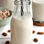 Garrafinha de vidro pequena com leite vegetal de amêndoas e um canudo preto e branco listrado, ao fundo mais leite dentro de vidros e o resíduo do leite. Na mesa branca algumas amêndoas espalhadas