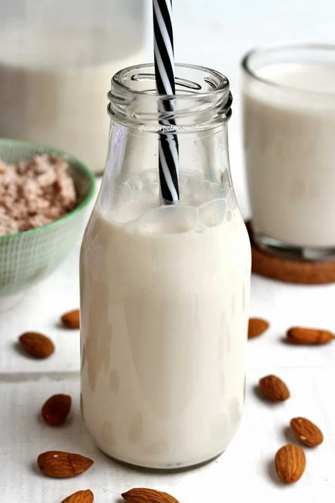 Pequeña botella de vidrio con leche de almendras vegetal y una pajita a rayas blancas y negras, al fondo más leche en vasos y los residuos de leche. Sobre la mesa blanca algunas almendras esparcidas
