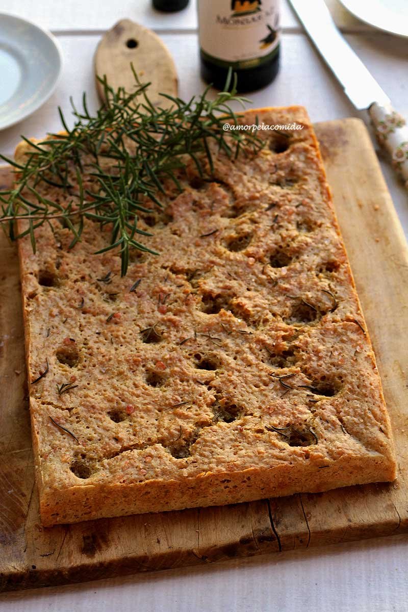 Pão focaccia retangular com furinhos na superfície, um ramo de alecrim por cima decorando sobre uma tábua de madeira em uma mesa branca