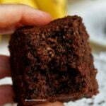 Mão segurando fatia de bolo de banana com chocolate mordido mostrando a textura aerada, ao fundo bananas desfocadas