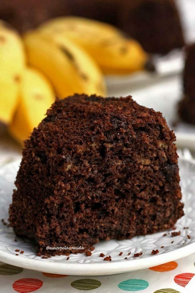 Fatia de bolo de banana com chocolate sobre prato redondo pequeno em uma mesa branca, ao fundo uma penca de banana desfocada e atrás o bolo partido