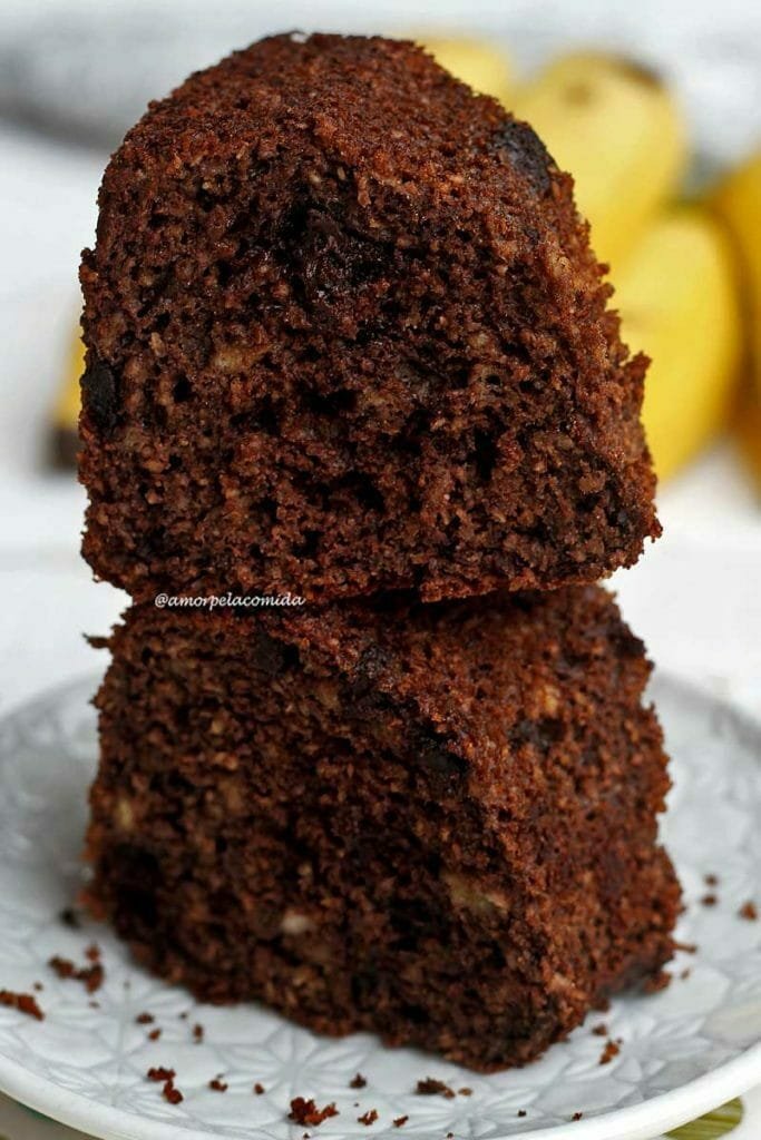 Duas fatias de bolo de banana com chocolate empilhadas em um prato redondo pequeno em uma mesa branca, ao fundo bananas desfocadas