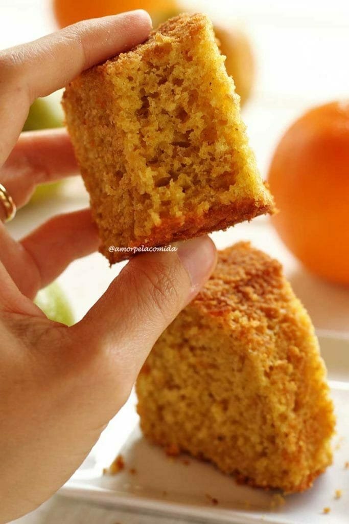 Mão segurando pedaço de bolo de laranja mordido mostrando a textura aerada da massa, abaixo uma fatia de bolo sobre um prato quadrado branco pequeno e laranjas desfocadas sobre uma mesa branca