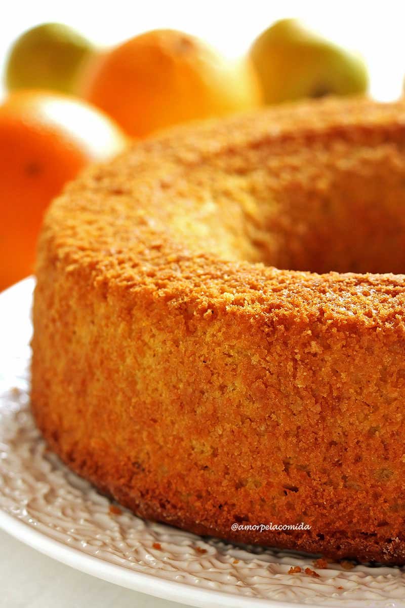 Bolo de laranja redondo com furo no meio sobre prato branco com a borda em relevo decorativo, ao fundo laranjas desfocadas em uma mesa branca