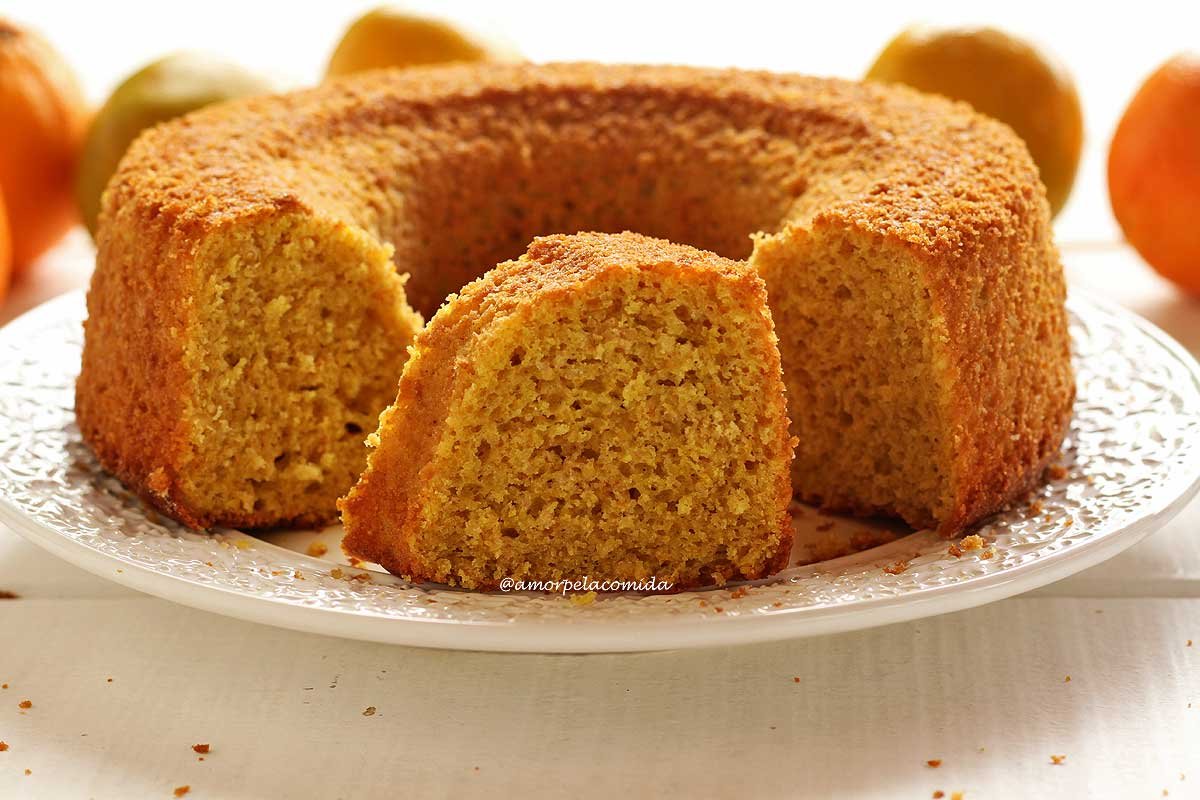 Bolo de laranja redondo com furo no meio partido sobre um prato branco, no meio do bolo tem uma fatia mostrando a textura aerada da massa, ao fundo laranjas desfocadas em uma mesa branca