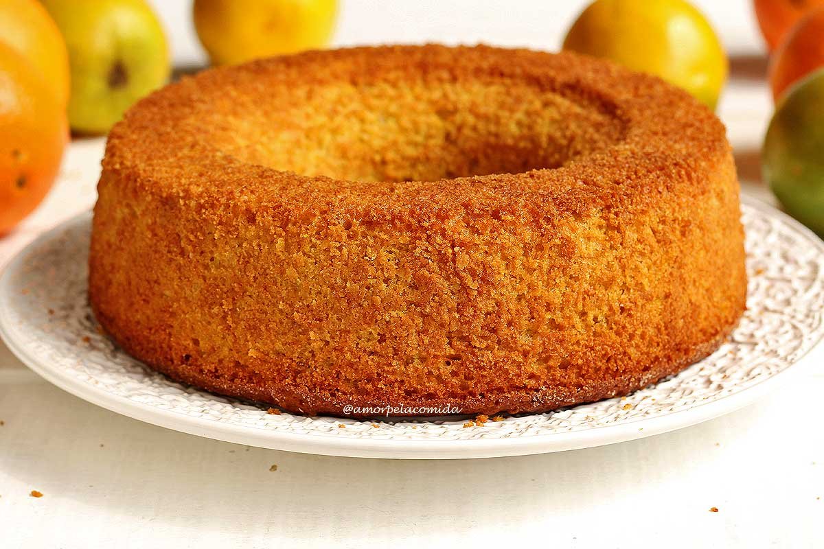 Bolo de laranja redondo com furo no meio sobre prato branco em uma mesa branca com laranjas desfocadas ao fundo