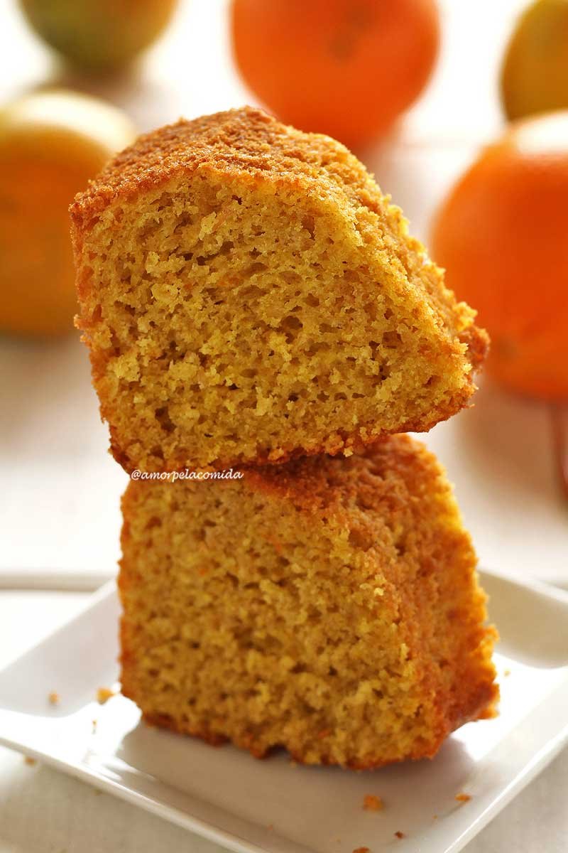 Duas fatias de bolo de laranja empilhadas sobre um prato quadrado branco pequeno em uma mesa branca com laranjas desfocadas ao fundo