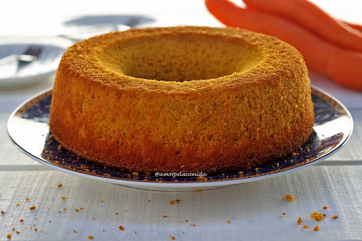 Bolo amarelo redondo com furo no meio sobre prato azul com detalhes amarelos em uma mesa branca, ao fundo cenouras desfocadas e dois pratos