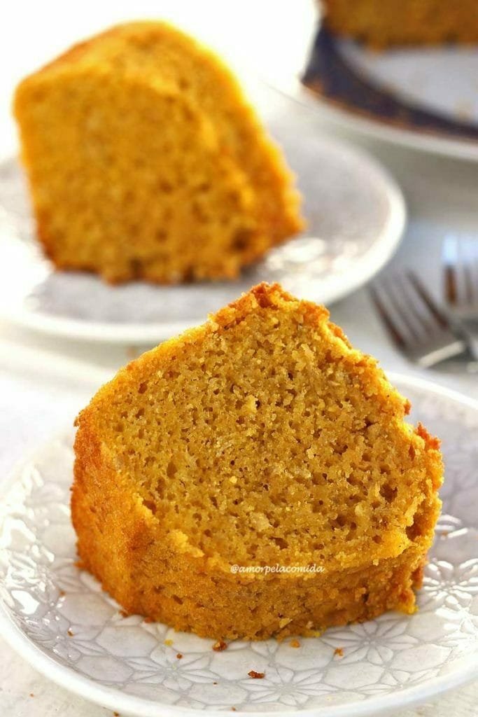 Duas fatias de bolo de cenoura com fubá sobre um prato redondo pequeno com detalhes geométricos em uma mesa branca